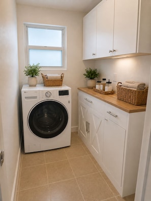A laundry room with a front-loading washing machine, cabinets, a window, and a countertop with baskets and toiletries.