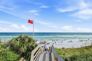 A beach with a wooden walkway leading to the shore, a red flag, and people enjoying the beach. - Sunnyside - 30a Blue Mountain Beach -Golf Cart or Walk to Beach With Ease (Santa Rosa Beach)