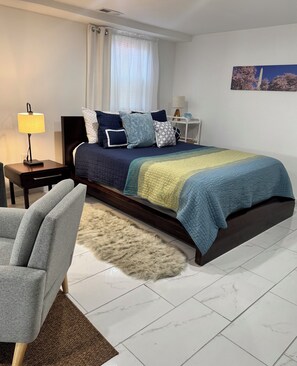 Ein Schlafzimmer mit Bett, Sessel, Lampe, Nachttisch, einem Fenster mit Vorhängen und einem Bild an der Wand.