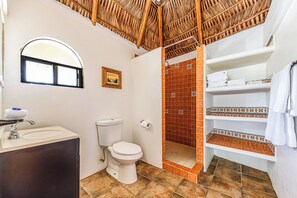 Casa de banho com sanita, lavatório, chuveiro e estantes com toalhas.