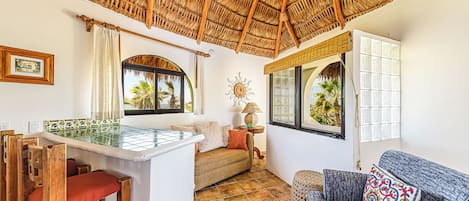 A room with a thatched roof, a tiled pool, a sofa, and a window with a view of palm trees.