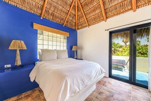 A bedroom with a blue wall, a bed with white bedding, two bedside lamps, a window with a decorative panel, and a view of a garden through a glass door. - Casita Del Mar - Steps from San Pedrito Surf Break (el Pescadero)