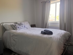Een netjes opgemaakt bed met witte lakens en een decoratief kussen, een opgerolde handdoek op het bed, en een raam met gordijnen.