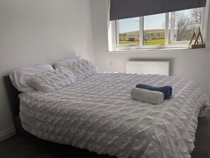 Een net opgemaakt bed met een witte dekbedovertrek en kussens, een raam met uitzicht op het buitengebied, en een radiator onder het raam.