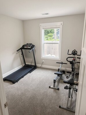 Ein kleiner Raum mit Teppichboden, ausgestattet mit Laufband, Ergometer und einem Fenster mit Blick auf ein Haus und Bäume.