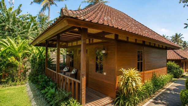 A wooden bungalow with a tiled roof, a porch with seating, and lush tropical vegetation. - Ubud 1BR Villa Wooden Resort Sahadewa (Gianyar)