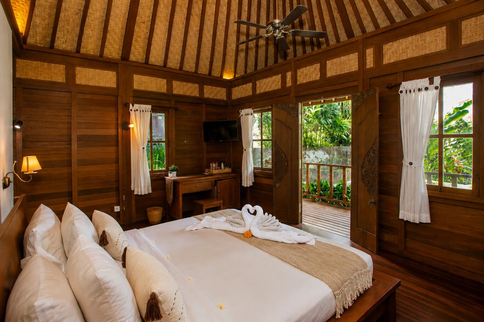 A wooden room with a bed, a desk, and a TV. There are windows with curtains and a view of greenery outside.