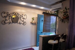 Ein Zimmer mit blauer Tür, einem Zierbild, einer kleinen Pflanze, einem Fenster mit Jalousien und einem Sessel.