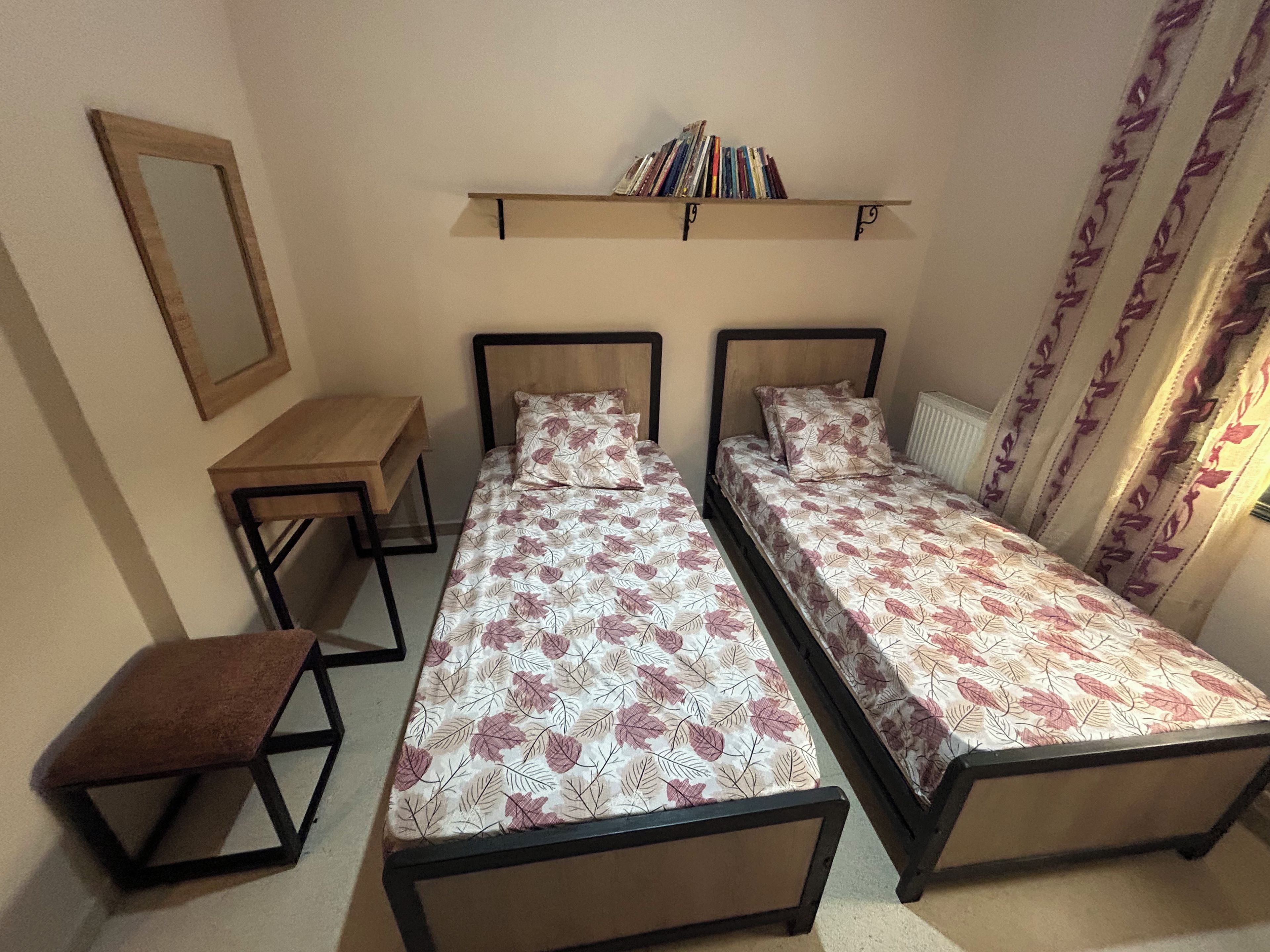 Quarto com duas camas de solteiro, uma mesinha, um espelho e uma estante com livros.