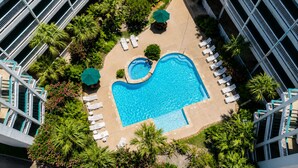 Ein weiter Blick von oben auf einen Hotelpoolbereich mit einem rechteckigen Pool, umgeben von Liegestühlen und Sonnenschirmen, mit Palmen und einem Gebäude im Hintergrund.