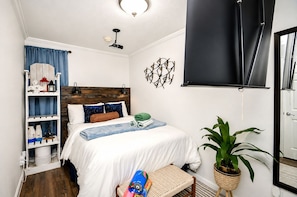 Un dormitorio con cama, repisas con objetos decorativos, una planta en maceta y una ventana con persiana.