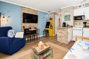 Ein Wohnzimmer mit blauem Sofa, Couchtisch, Fernseher und einer kleinen Küchenzeile mit Mikrowelle und Herd.