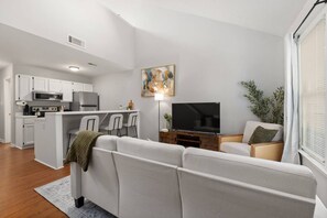 Ein modernes Wohnzimmer mit einer Couch, einem Fernseher, einem Essbereich mit Stühlen und einer Küche mit weißen Schränken.