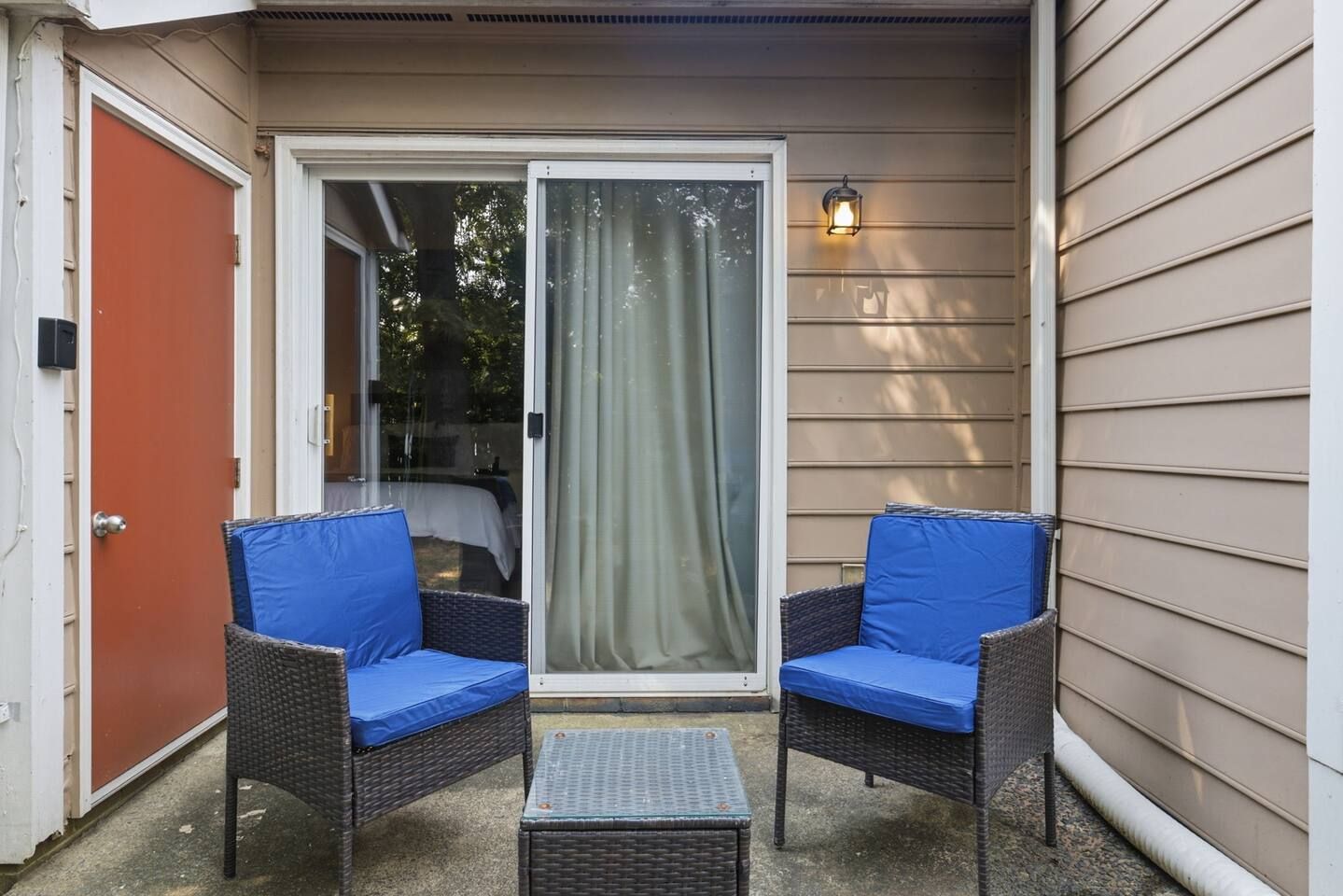 Un patio con dos sillas de mimbre, una mesita y una puerta corrediza de vidrio, además de un aplique de pared.