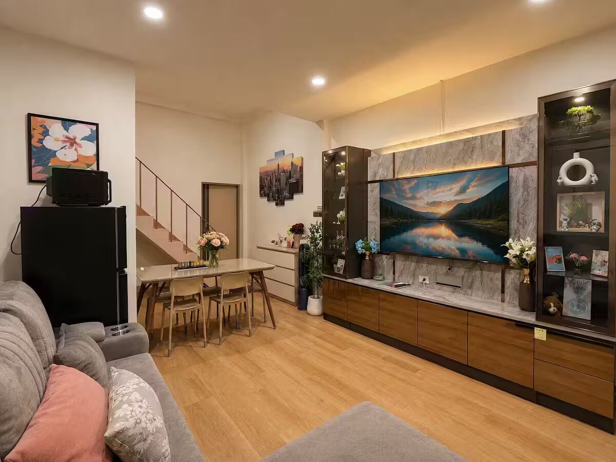 A modern living room with a grey sofa, a dining area with a table and chairs, a kitchen with a large TV, and a staircase.