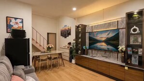 Een moderne woonkamer met een grijze bank, een eethoek met een tafel en stoelen, een keuken met een grote televisie, en een trap.