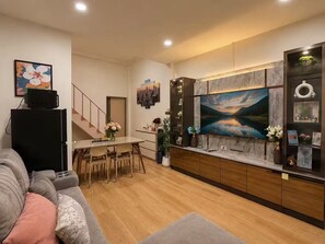 A modern living room with a grey sofa, a dining area with a table and chairs, a kitchen with a large TV, and a staircase. - Entire home in Ratchathewi, Thailand (Thanon Petchaburi, Ratchathewi)