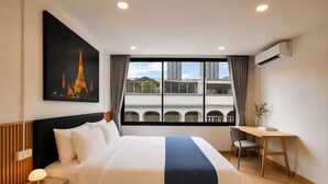 Een hotelkamer met een bed, nachtkastje, bureau, stoel en een groot raam met uitzicht op gebouwen.