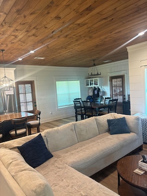 Una sala de estar amplia con techo de madera, un sofá grande color beige y un comedor con mesa y sillas de madera.