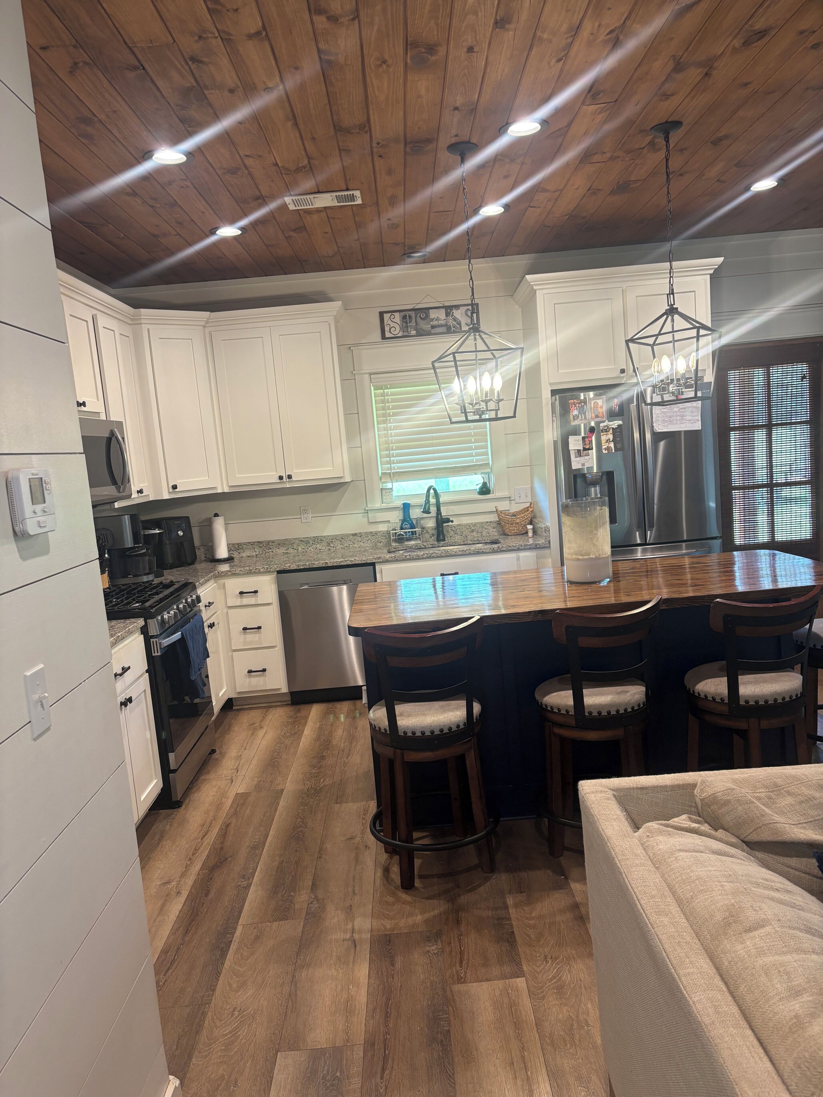 A kitchen with white cabinets, a stainless steel refrigerator, a wooden ceiling, and a bar area with stools.