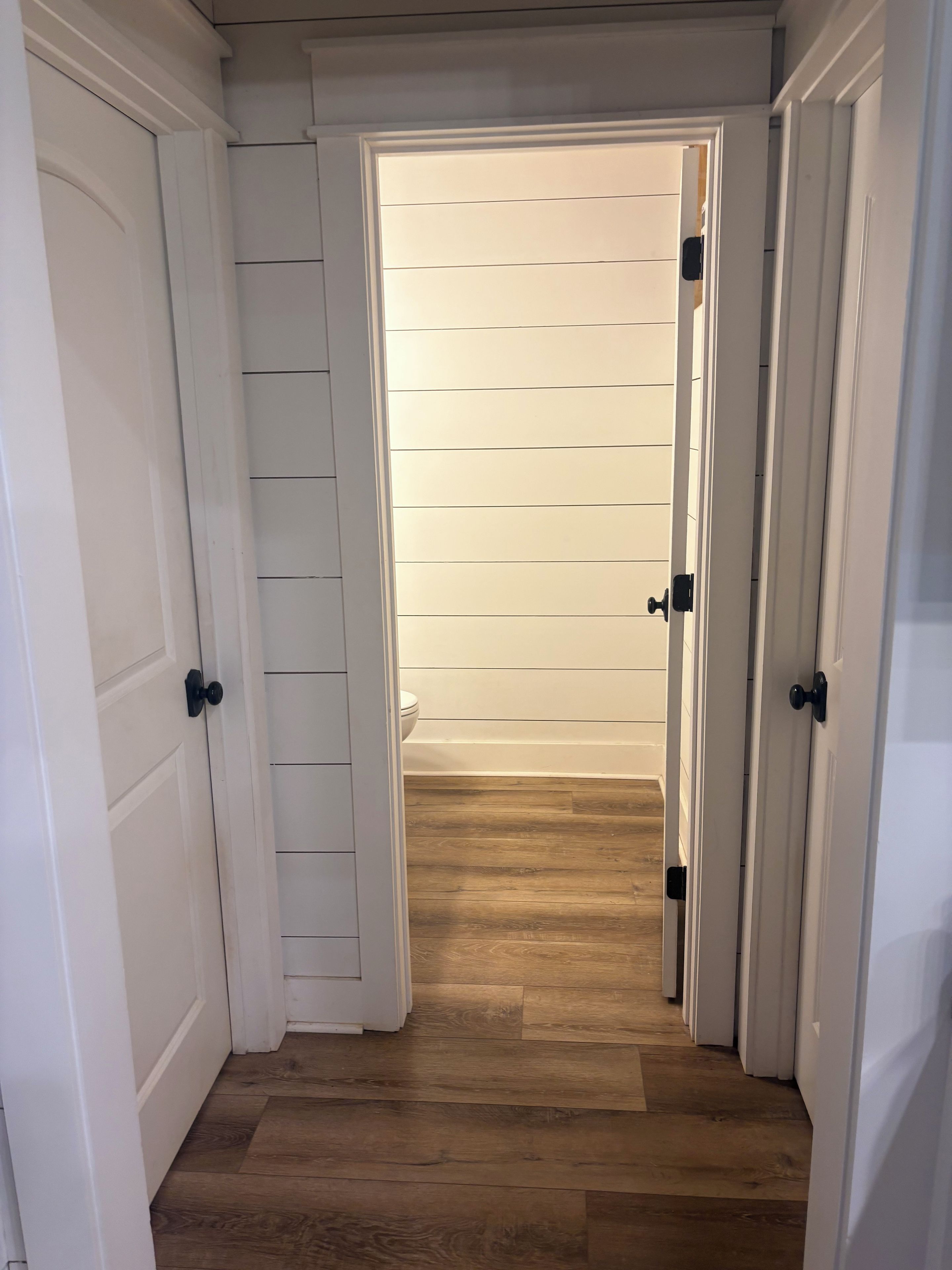 Un couloir aux portes blanches et au plancher en bois mène à une pièce aux murs recouverts de lambris horizontaux.