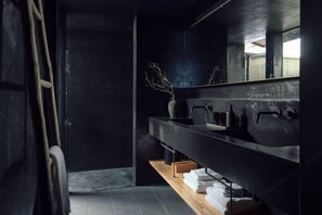 Une salle de bain moderne aux couleurs sombres, dotée d’un grand miroir et d’une étagère en bois où sont posées des serviettes.