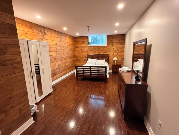A bedroom with a wooden floor, a bed, a dresser, a mirror, and a window.