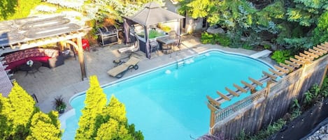 A swimming pool with a wooden deck and lounge chairs, surrounded by trees and a covered seating area.
