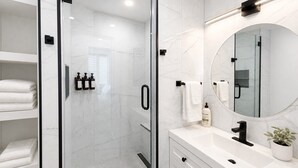 A modern bathroom with a glass-enclosed shower, white marble walls, a round mirror, and a white countertop with a sink and a potted plant. - 3 bedroom downtown condo - sleeps 6 - lake & pools (Kelowna)
