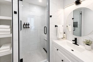 Un baño moderno con ducha envuelta en vidrio, paredes de mármol blanco, un espejo redondo y una encimera blanca con un lavamanos y una planta en maceta.