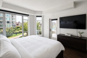 Un dormitorio con una cama grande, televisión, cómoda oscura y puerta corrediza de vidrio que da a un balcón con vista a una piscina y edificios.