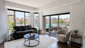 A modern living room with a sofa, coffee table, and a view of a swimming pool and buildings. - 3 bedroom downtown condo - sleeps 6 - lake & pools (Kelowna)