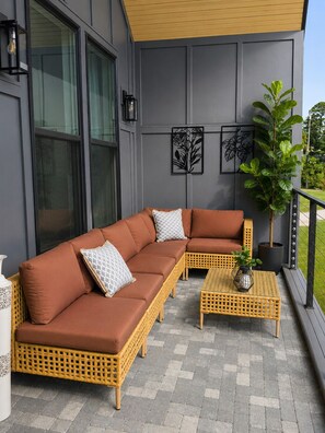 Un área de estar exterior moderna con un sofá marrón y dos mesas de mimbre, una planta en maceta y decoración en la pared.