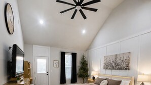 Ein Schlafzimmer mit Bett, Nachttischen, einem Fernseher, einem Deckenventilator, einem Fenster mit Vorhängen und einem an der Wand befestigten Kunstwerk.