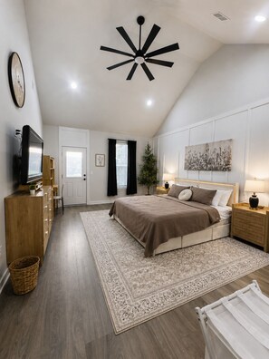A bedroom with a bed, bedside tables, a television, a ceiling fan, a window with curtains, and a wall-mounted artwork.
