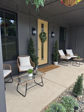 A modern outdoor seating area with wicker chairs and a glass table, a door with a wreath, and a welcome mat.