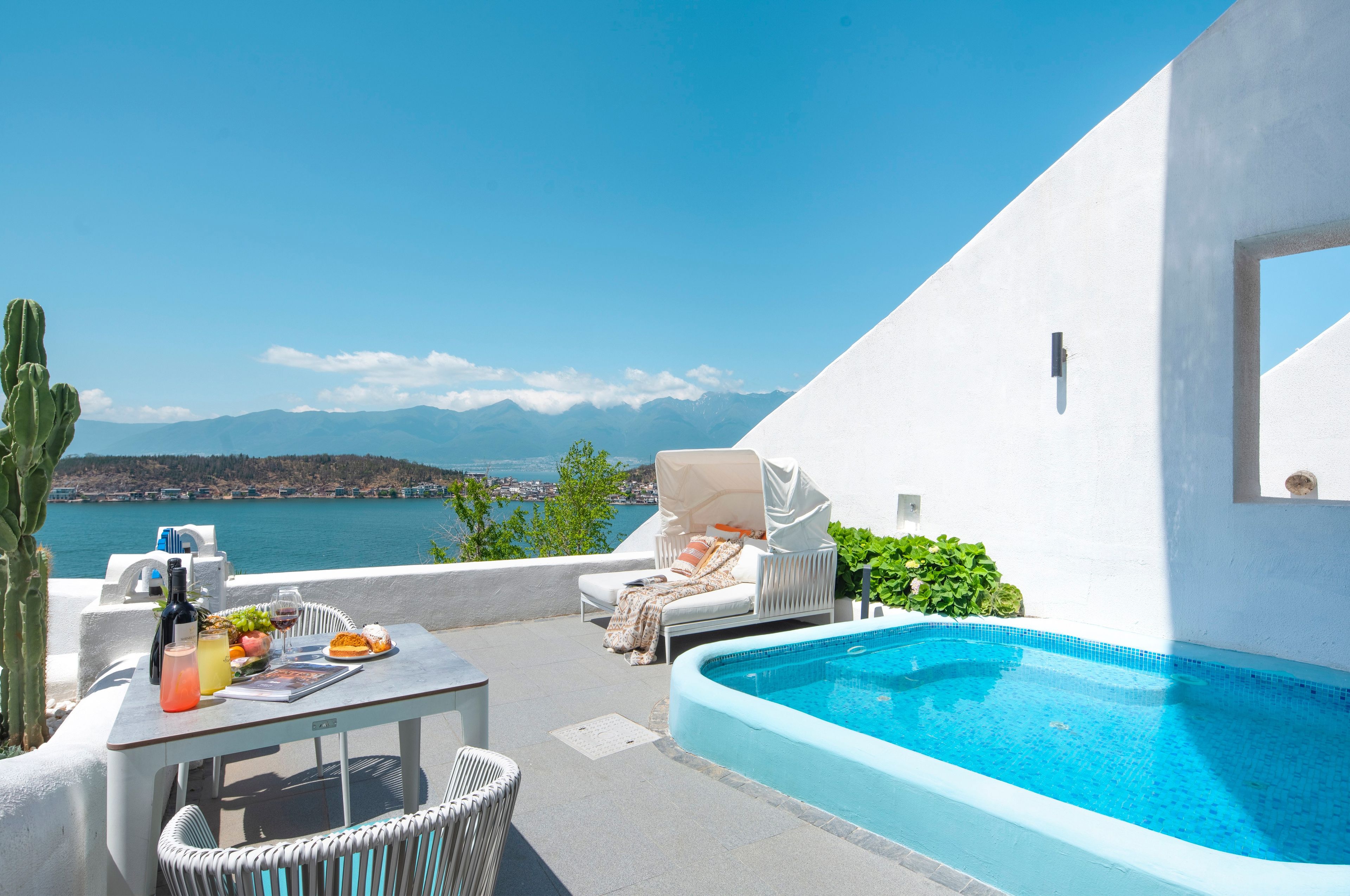 Zona de piscina con vista al mar, una mesa con bebidas y comida, y una silla lounge cubierta.