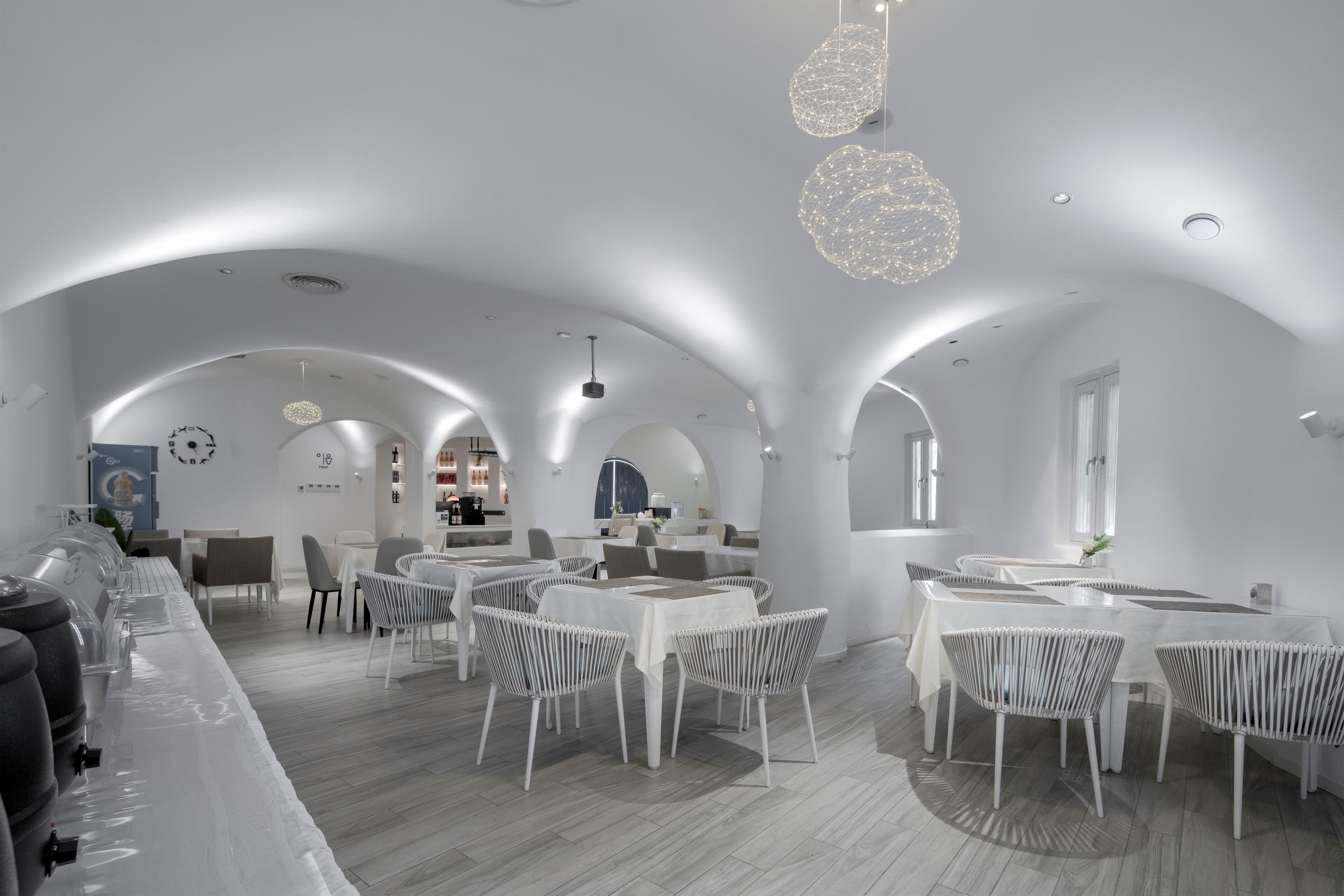 Un comedor moderno con mesas y sillas blancas, techos abovedados e iluminación colgante.