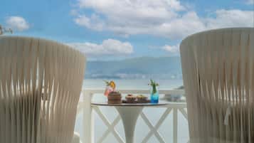 Un balcón con dos sillas blancas, una mesita con una bebida y un postre, y vistas al mar y las montañas.