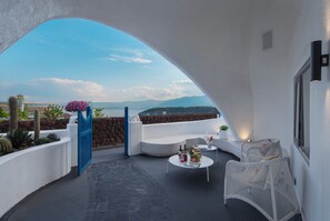 Un balcón con pared blanca, un portón azul, un sofá blanco, una mesa redonda con bebidas, y vista al mar y las montañas.