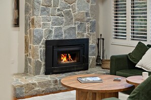 A stone fireplace with a fire, a green sofa, and a round wooden table with books. - Hallia House – Coastal Retreat (Readhead)