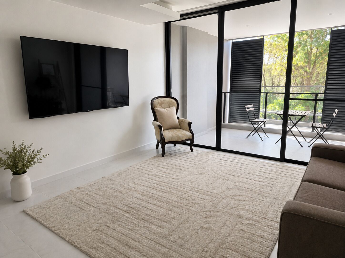 A modern living room with a large flat-screen TV, a comfortable armchair, a beige rug, and a view of an outdoor patio with a table and chairs.