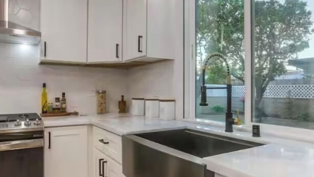 Cocina moderna con muebles blancos, estufa de acero inoxidable, lavavajillas y un amplio ventanal con vistas a árboles.