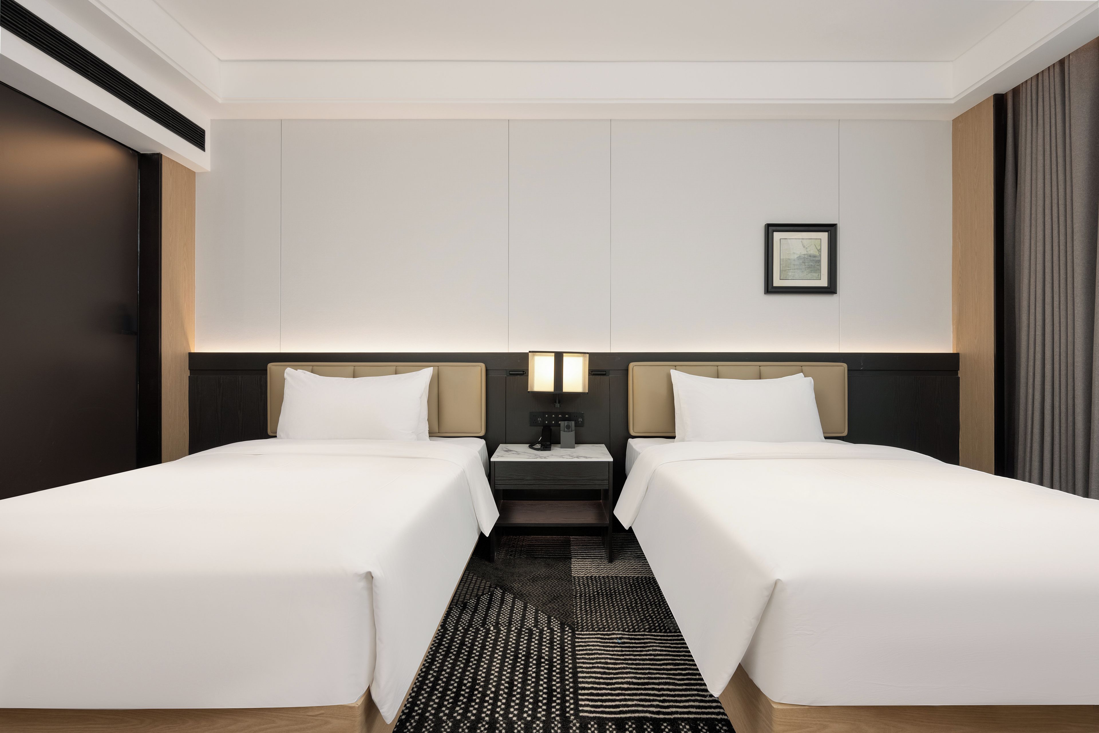 Una habitación de hotel con dos camas individuales, una mesita de noche con lámpara, un cuadro en la pared y una alfombra estampada.