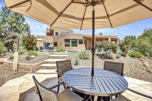 Une terrasse avec une table ronde et des chaises, un parasol beige et, au fond, une maison moderne.