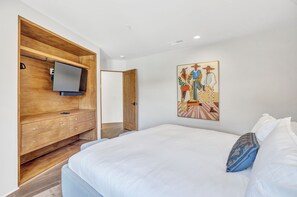 Quarto moderno com uma cama grande, penteadeira de madeira com uma TV de tela plana, porta que dá para outro cômodo e um quadro na parede.