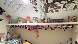 A room with a vintage chair, a desk with a lamp, a shelf with various items, and a wall with a tree and cat illustration.