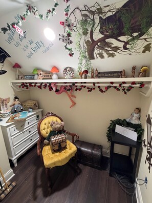 A room with a vintage chair, a desk with a lamp, a shelf with various items, and a wall with a tree and cat illustration.