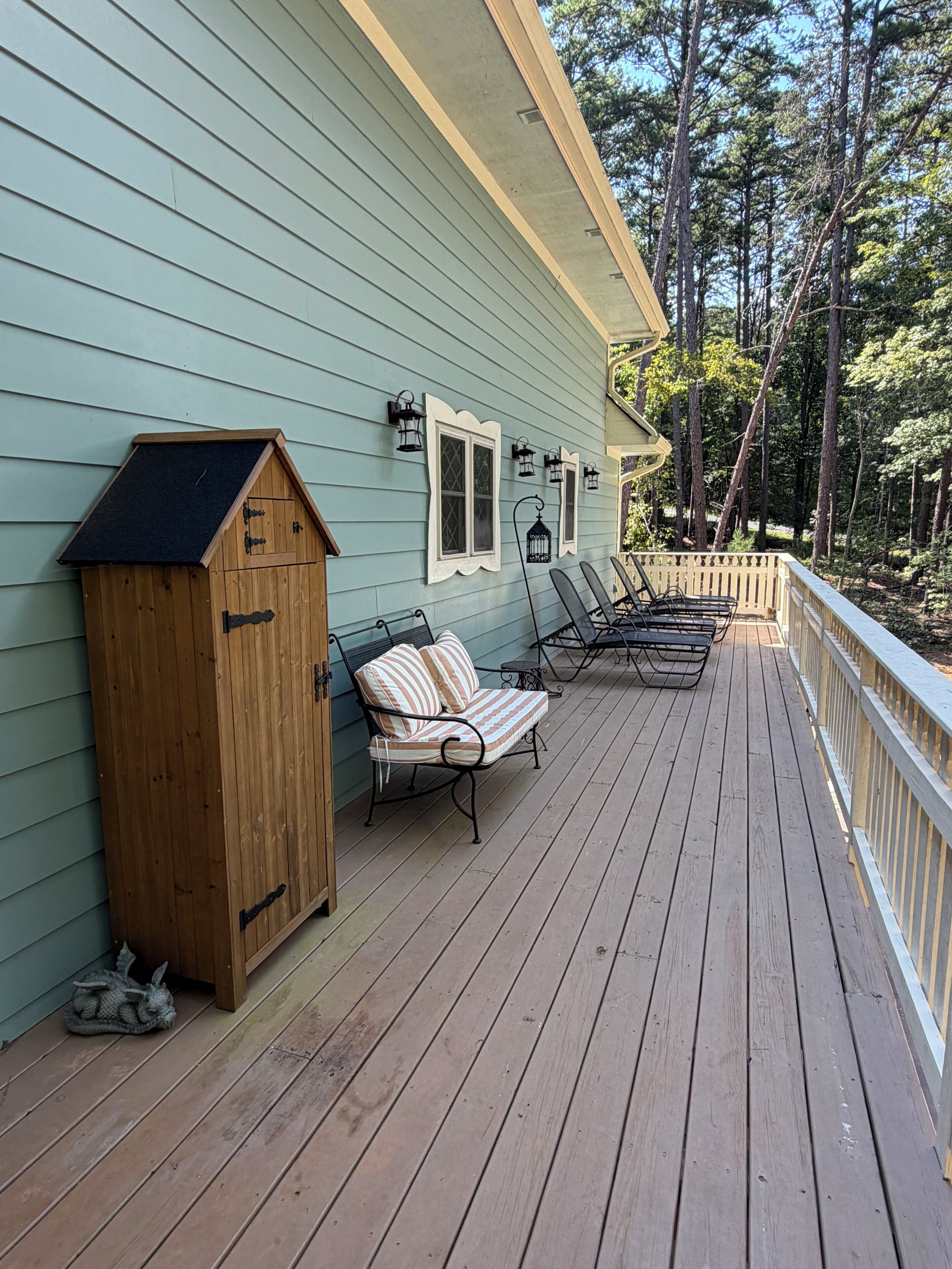 Une terrasse en bois avec un banc intégré et une petite construction en bois, probablement un abri de jardin, située à côté d’une maison à l’extérieur bleu clair.