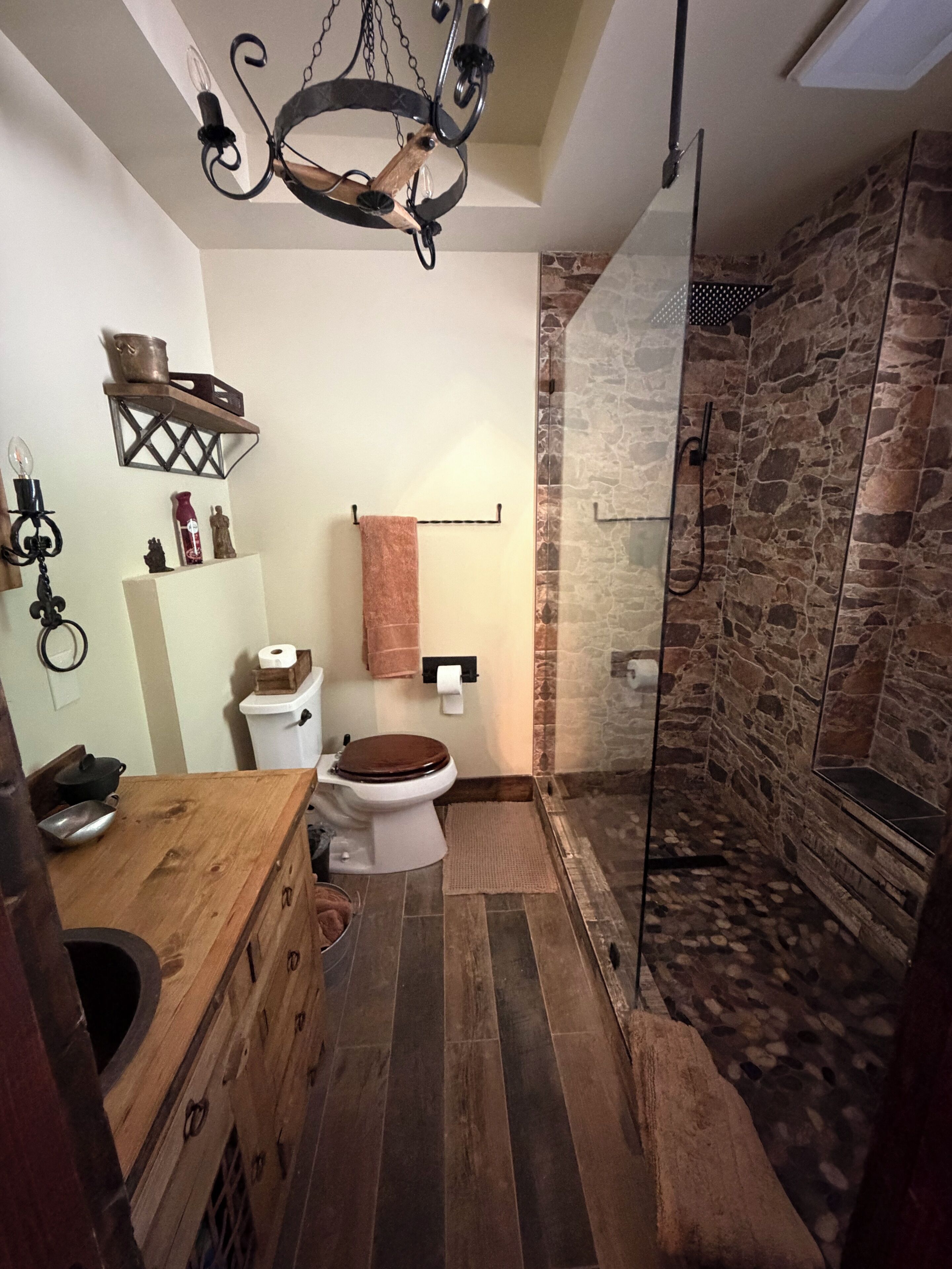 A bathroom with a stone shower wall, a wooden vanity, and a toilet with a brown seat.